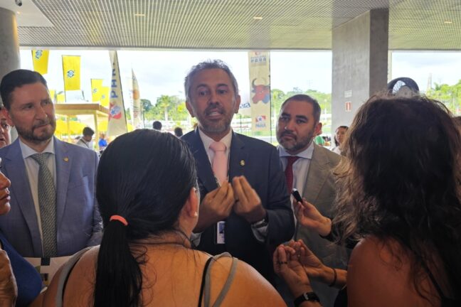 Foto de um grupo de pessoas reunidas em pé, em um ambiente amplo e iluminado, enquanto um homem de terno escuro e gravata rosa, ao centro, fala com gestos firmes das mãos, como quem explica ou responde a demandas. Ele está cercado por mulheres e homens que o escutam atentamente, alguns segurando celulares para registrar a conversa. À esquerda e à direita, outros dois homens de terno acompanham a cena com expressões sérias. Ao fundo, do lado externo, aparecem bandeiras, árvores e estruturas da COP30