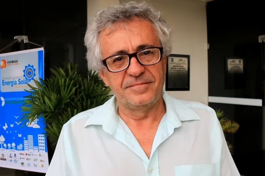 A imagem mostra um homem de cabelos grisalhos e curtos, usando óculos de armação escura e uma camisa social clara. Ele está de pé em um ambiente externo, próximo a uma entrada de prédio. Atrás dele há plantas e um banner azul com a palavra “Energia Solar” e ícones de sol, prédios e nuvens