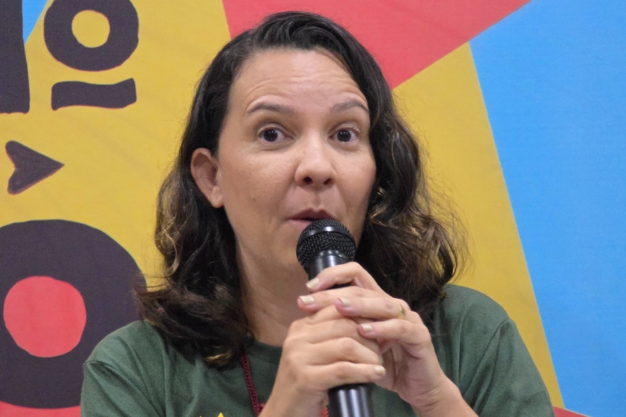 Foto de mulher falando ao microfone, em um ambiente de evento ou debate. Ela tem pele clara, cabelos castanhos ondulados na altura dos ombros e veste uma camiseta verde. Seu olhar está atento e ligeiramente surpreendido, como se estivesse reagindo a algo dito ou preparando uma resposta. O fundo é colorido, composto por formas geométricas grandes em tons de amarelo, azul e vermelho
