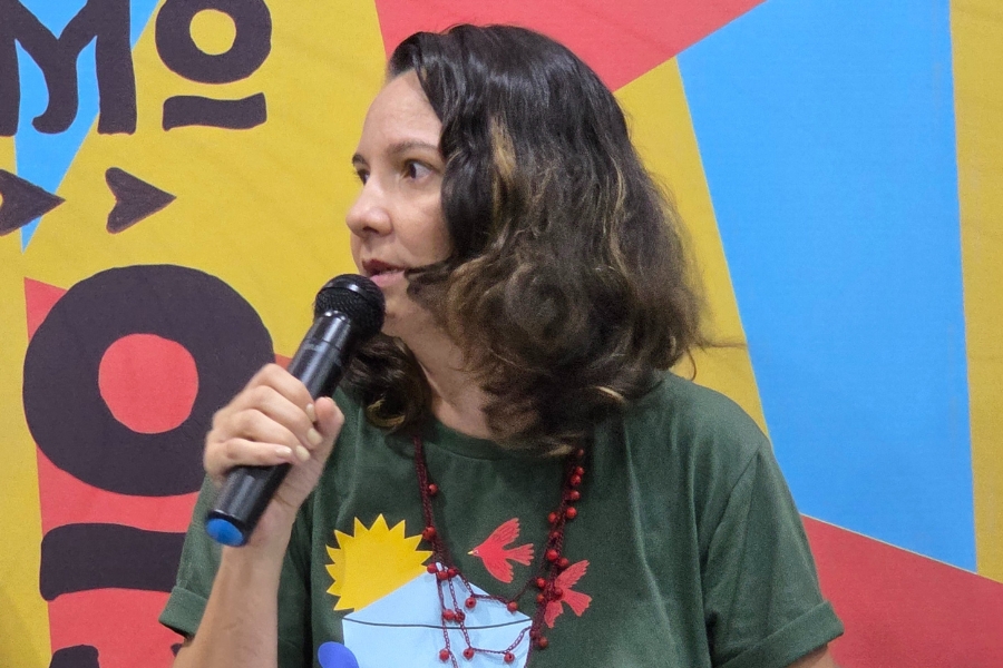 Foto de mulher segurando um microfone enquanto olha para a esquerda, como se estivesse respondendo a alguém ou acompanhando uma fala. Ela tem pele clara, cabelos castanhos ondulados na altura dos ombros e usa uma camiseta verde estampada com ilustrações coloridas, além de um colar vermelho. O fundo é vibrante, composto por grandes formas geométricas em amarelo, azul e vermelho