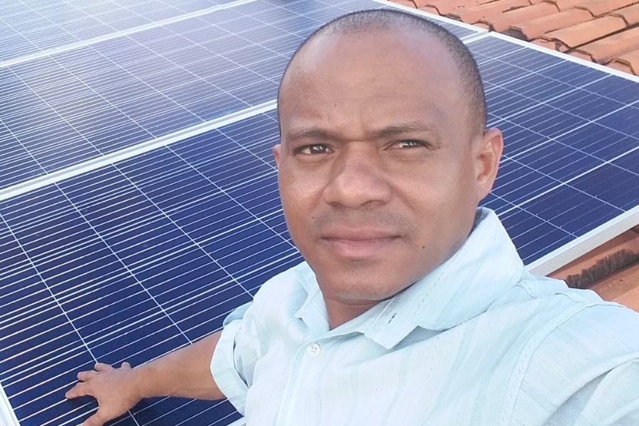 A imagem mostra um homem negro calvo, vestindo uma camisa clara, posando para uma selfie ao ar livre. Ele está em frente a um conjunto de painéis solares instalados sobre um telhado de telhas alaranjadas
