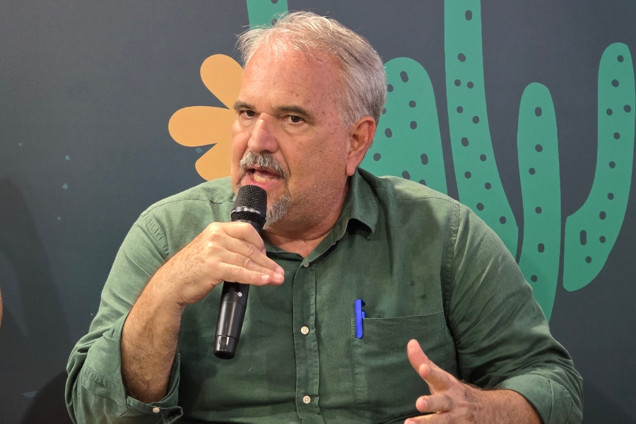 A foto mostra um homem de meia-idade, de pele clara e cabelos grisalhos, sentado e falando ao microfone. Ele veste uma camisa verde de mangas compridas, ligeiramente enrugada, com uma caneta azul no bolso. Sua expressão é séria e concentrada, enquanto gesticula com a mão livre para enfatizar o que diz. Ao fundo, há um painel escuro com ilustrações estilizadas em verde e laranja que lembram formas vegetais