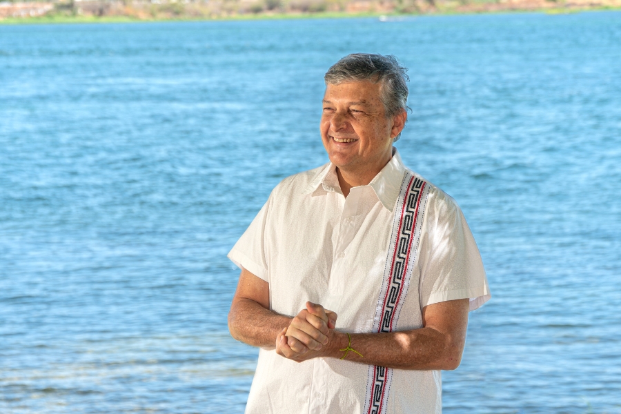 Na imagem, um homem está de pé à beira de um rio, com a água azul ao fundo e uma faixa de vegetação distante na margem oposta. Ele veste uma camisa branca de manga curta com um bordado colorido em uma das laterais e sorri levemente, olhando para o lado