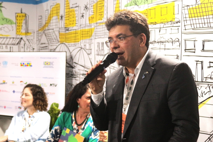 Foto um homem de terno escuro e camisa clara falando ao microfone durante um evento. Ele usa óculos e crachá no pescoço. Atrás dele, duas mulheres estão sentadas, uma de blusa azul e outra verde, observando a fala. O fundo é composto por um painel ilustrado com desenhos de prédios, árvores e estruturas urbanas em cores vivas