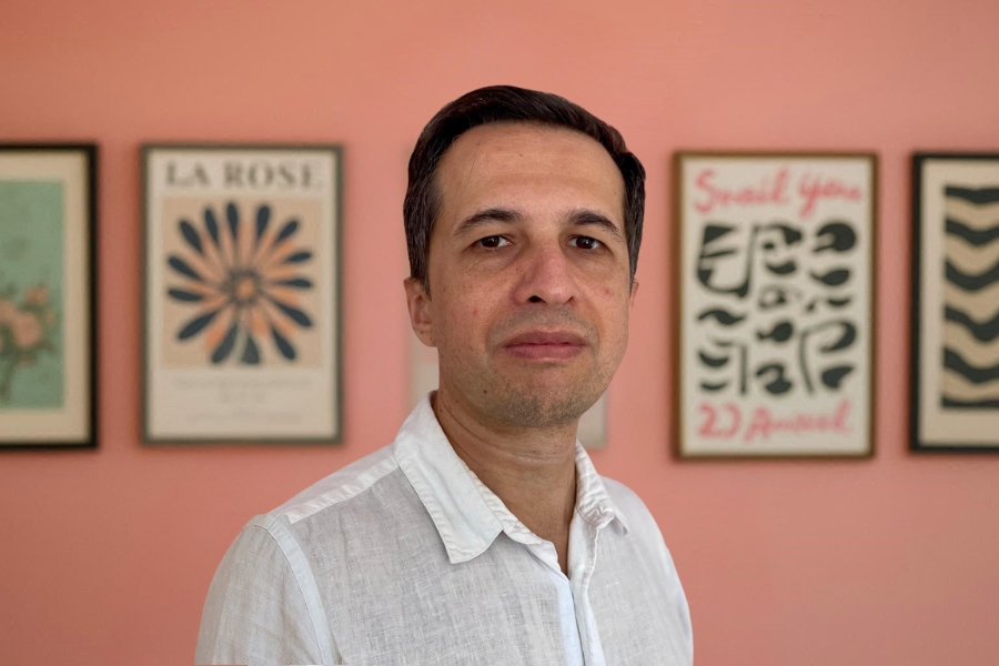 Foto um homem de meia-idade com expressão neutra, cabelos curtos escuros e leves sinais de barba, posicionado em primeiro plano e olhando diretamente para a câmera. Ele veste uma camisa branca de linho e está em um ambiente interno com parede rosa ao fundo, decorada com quadros emoldurados com ilustrações e tipografias artísticas, levemente desfocados