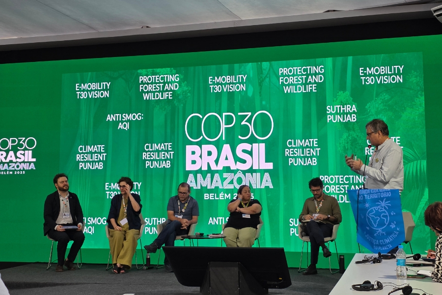 Foto de uma mesa de debate diante de um grande painel verde com as palavras “COP30 Brasil Amazônia – Belém 2025” ao centro e diversos temas climáticos espalhados ao redor. Seis pessoas participam da cena: cinco estão sentadas à esquerda, formando uma fileira, enquanto uma pessoa à direita está de pé, falando ao microfone e segurando uma bandeira azul dobrada. Os participantes usam crachás e roupas casuais. À frente deles há uma mesa baixa com garrafas e materiais de apoio