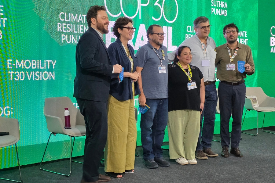 Um grupo de seis pessoas posa sorrindo sobre um palco iluminado por um grande painel verde com a inscrição “COP30 BRASIL”. Elas estão lado a lado, em pé, algumas segurando pequenos objetos azulados, como brindes ou placas. No grupo, há três mulheres e três homens, todos usando crachás pendurados no pescoço. As roupas são sociais-casuais, com tons que variam do preto ao bege, passando por azul e verde. Atrás deles, cadeiras modernas e uma garrafa rosa sobre um assento compõem o cenário de um espaço de debate