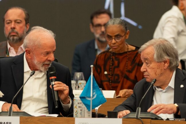 Fotografia mostra duas pessoas em destaque sentadas lado a lado à mesa principal, em um ambiente de conferência. À esquerda, um homem de barba branca fala ao microfone e gesticula levemente, voltado para outro homem grisalho, que o escuta atentamente. Entre eles há bandeiras pequenas sobre a mesa, incluindo uma da Organização das Nações Unidas (ONU). Ao fundo, outras pessoas acompanham a conversa, entre elas uma mulher de óculos e colar escuro