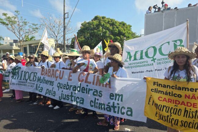 A foto, tirada em plano médio em um dia ensolarado, mostra um grupo de mulheres, a maioria usando chapéus de palha e camisetas brancas, participando de uma manifestação de rua. Elas estão alinhadas e seguram duas faixas principais. A faixa central, grande e branca com letras verdes e vermelhas, diz em destaque: "BABAÇU E CLIMA. Mulheres quebradeiras em luta pela justiça climática". À frente, no canto inferior direito, uma mulher segura uma faixa amarela que diz: "Financiamento climático NÃO é CARIDADE. É reparação histórica e justiça social". O logotipo do MIQCB (Movimento Interestadual das Quebradeiras de Coco Babaçu) aparece em algumas bandeiras e faixas, incluindo uma grande bandeira branca ao fundo, que identifica o movimento nos estados do Maranhão, Pará, Piauí e Tocantins. A manifestação acontece em uma rua pavimentada, com vegetação, árvores e edifícios ao fundo, e algumas pessoas assistindo de cima de uma estrutura à direita