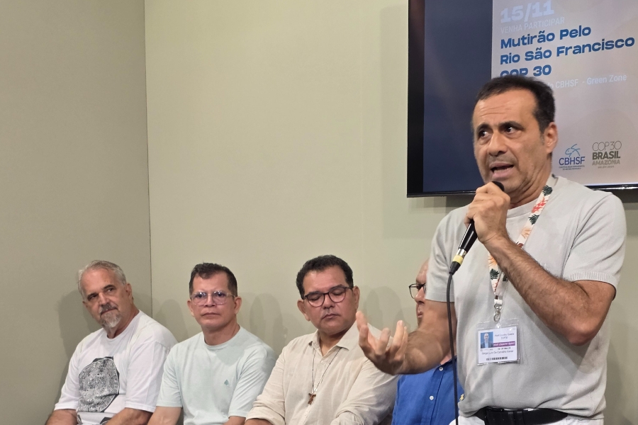 A foto de ambiente interno captura um momento em um evento, com quatro homens sentados e um em pé contra uma parede lisa de cor cinza-claro. Em primeiro plano, no lado direito, um homem de pele morena, vestindo uma camisa cinza-claro e um crachá de identificação, está em pé e falando com emoção em um microfone que segura com a mão esquerda, enquanto gesticula com a mão direita aberta. O olhar dele está direcionado ligeiramente para a frente e para a esquerda. Atrás dele, à esquerda, quatro outros homens estão sentados em uma fila, prestando atenção: o primeiro da esquerda é um homem grisalho de barba vestindo uma camiseta branca; os dois seguintes são homens de meia-idade, ambos usando camisas de cores claras (verde-água e bege), com óculos, olhando para frente; e o último, apenas parcialmente visível, parece estar de óculos. No canto superior direito, há uma tela de televisão ou monitor que exibe texto, incluindo a data "15/11" e as frases "MUTIRÃO PELO RIO SÃO FRANCISCO" e "COP 30 - Green Zone". A iluminação é uniforme e clara