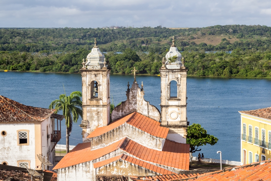 Foto de  uma vista de cima de uma paisagem histórica e natural, focada no telhado e nas torres de uma igreja antiga no centro. O edifício religioso apresenta um telhado de telhas de barro de cor laranja avermelhada desgastadas. De trás do telhado, erguem-se duas imponentes torres sineiras em estilo colonial, de cor clara, quase branca, com detalhes em pedra cinza escura, cada uma com aberturas em arco para os sinos (o sino é visível na torre esquerda) e encimadas por pináculos. Entre as torres, há uma cruz simples. Em primeiro plano e nas laterais, podem-se ver outros telhados de casas históricas com as mesmas telhas de barro e paredes claras (amarelas e brancas) com janelas escuras e varandas. Uma palmeira esguia se destaca à esquerda. A característica mais marcante do fundo é o vasto e azulado Rio São Francisco, que domina o plano médio. A margem oposta do rio é coberta por uma densa e exuberante vegetação verde, que se estende até o horizonte sob um céu claro com algumas nuvens dispersas