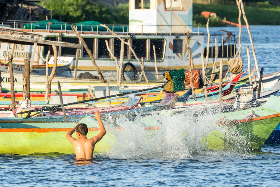 A foto mostra uma cena vibrante e animada em um corpo d'água. Em primeiro plano, um menino de costas, sem camisa, está brincando na água, batendo nela com as mãos levantadas e criando um grande e vigoroso respingo que se espalha sobre a superfície. A água ao redor é calma e azul-escura. O garoto está perto de uma embarcação de cor verde limão e turquesa. Ao fundo, uma série de barcos de pesca coloridos (em tons de amarelo, vermelho e branco, entre outros) estão amarrados e agrupados, preenchendo o plano médio. Por trás deles, emerge uma estrutura de dois andares com uma pequena janela no topo, que se parece com a cabine de um barco maior com algumas formas geométricas pintadas