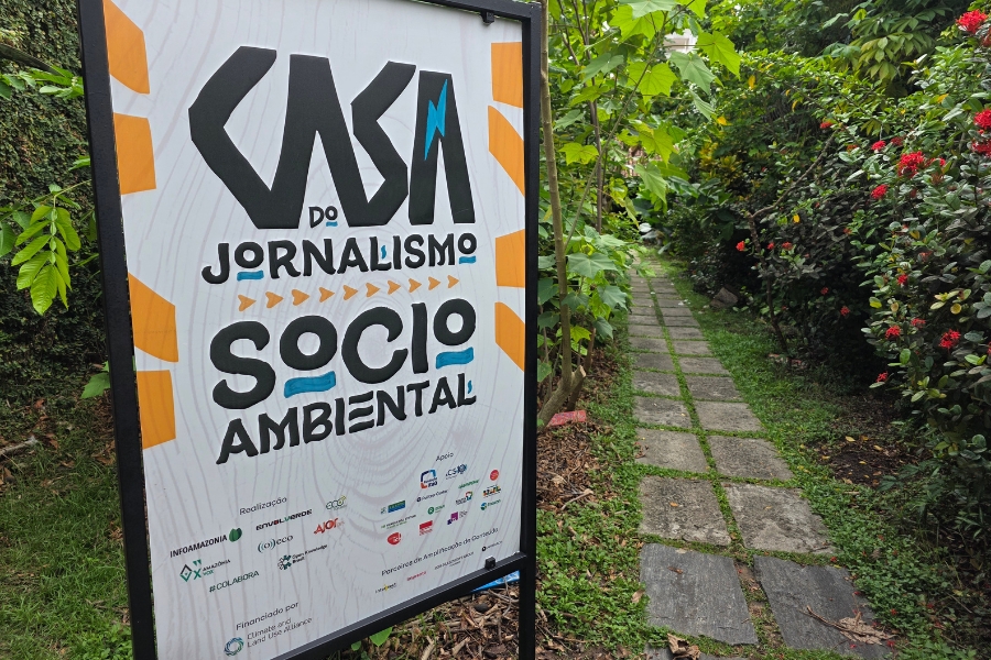 A foto em primeiro plano captura uma placa de sinalização vertical com fundo branco, centralizada na imagem e emoldurada em preto, indicando a "CASA DO JORNALISMO SOCIOAMBIENTAL" em letras pretas e azuis escuras grandes, estilizadas e impactantes. Abaixo do título principal, em letras menores, a placa lista diversas instituições parceiras e apoiadoras, incluindo logotipos coloridos, e informa que o projeto é financiado pelo Fundo Clima e Uso da Terra. A placa está posicionada em um ambiente externo e verdejante, à direita do quadro, revelando um caminho estreito e rústico de lajotas quadradas de cimento, ladeado por folhagens densas e plantas com flores vermelhas. A vegetação exuberante cria uma atmosfera de natureza preservada, sugerindo que o local da "Casa" está imerso em um jardim ou área arborizada.