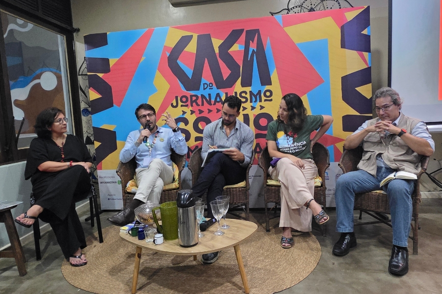 Foto de um ambiente interno e bem iluminado, exibe uma mesa redonda ou painel de discussão com cinco participantes sentados em cadeiras de vime ou material similar, dispostos em semicírculo voltados para frente. Da esquerda para a direita, estão: uma mulher de óculos e roupa escura, um homem de camisa social azul clara e calças cáqui falando em um microfone; um segundo homem de camisa social azul escura e calças pretas, olhando para baixo e escrevendo em um bloco; uma mulher vestindo uma camiseta verde escura estampada e calças claras; e, por último, um homem de colete cáqui, camisa azul clara e jeans, com óculos, apoiando o rosto na mão. No centro, à frente dos palestrantes, há uma pequena mesa redonda de madeira com alguns objetos, como copos, uma jarra e xícaras. O fundo é dominado por um grande painel ou banner com design gráfico vibrante nas cores vermelho, amarelo, azul e preto, com o logotipo estilizado e o texto "CASA DO JORNALISMO SOCIOAMBIENTAL"