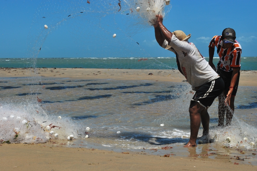 A foto mostra dois pescadores na beira de uma praia. O pescador em primeiro plano, usando uma camisa clara, shorts escuros e um chapéu de palha, está com os braços erguidos acima da cabeça, no meio do arremesso ou recolhimento de uma grande rede de pesca branca. A rede está aberta, com o peso do material e possivelmente da pesca, e várias boias brancas ovais estão visíveis, algumas ainda na rede e outras espalhadas na água rasa e na areia ao redor. O segundo pescador, à direita, usa uma camisa listrada em laranja e preto e um boné, e está curvado, segurando ou ajustando a rede. Eles estão na transição entre a areia clara da praia e a água do mar, que tem áreas mais escuras de algas ou lama. Ao fundo, o mar é calmo e se encontra com a linha do horizonte azul-claro, sob o céu igualmente azul