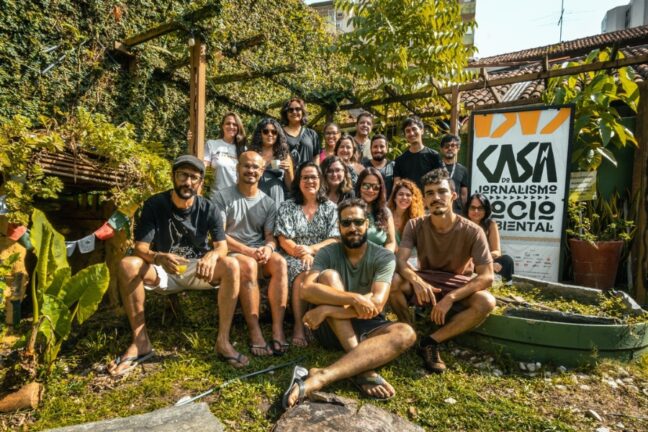Foto em plano médio, tirada ao ar livre em um dia ensolarado, mostra um grupo de cerca de dezesseis pessoas, incluindo homens e mulheres de diversas idades, reunidas em um ambiente que parece ser um jardim ou quintal. O grupo está sentado e em pé, olhando para a câmera e sorrindo. O fundo é composto por paredes e cercas cobertas por bastante vegetação verde, criando uma atmosfera natural e acolhedora. No canto superior direito, há uma grande faixa ou banner em cores laranja e branco com o logotipo e o texto "CASA DO JORNALISMO SOCIOAMBIENTAL", indicando o contexto do encontro. As pessoas estão vestindo roupas casuais, em sua maioria claras ou de cores escuras neutras, e a iluminação é forte, com áreas de sombra e luz solar
