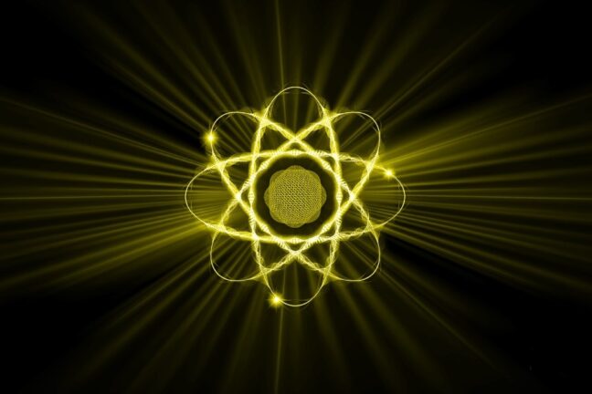 Esta imagem apresenta uma representação artística e estilizada de um átomo vibrante em tons de amarelo neon sobre um fundo preto sólido. No centro, há um núcleo esférico texturizado de onde partem diversas órbitas elípticas entrelaçadas, sugerindo o movimento de elétrons. O átomo inteiro parece brilhar intensamente, emitindo longos raios de luz amarela que se estendem em todas as direções até as bordas da imagem, criando um efeito visual de energia pulsante e radiante