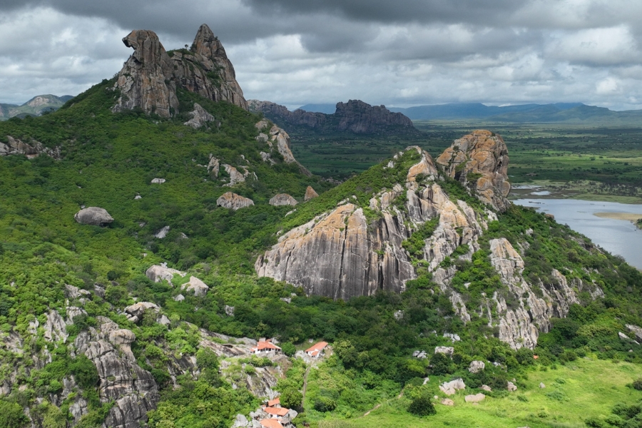 Foto de uma paisagem ampla dominada por grandes formações rochosas isoladas, com paredes íngremes e superfícies claras, emergindo de um manto verde de vegetação densa. Entre as rochas, a mata cobre encostas e vales, criando contraste entre o cinza das pedras e o verde intenso das árvores e arbustos. Ao fundo, o relevo se abre em planícies e serras mais distantes, sob um céu carregado de nuvens escuras, que anunciam chuva. Na parte inferior da cena, pequenas casas de telhado alaranjado aparecem encaixadas no pé das montanhas, reforçando a escala monumental das rochas e a integração entre presença humana e paisagem natural
