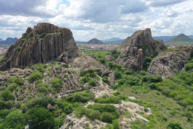 Fotografia aérea destaca duas imensas formações de rocha granítica escura e acinzentada que emergem verticalmente da paisagem. As rochas possuem texturas rugosas, com fendas profundas e marcas de erosão milenar, sendo parcialmente cobertas por uma vegetação rasteira e arbustos verdes que crescem em suas encostas e na base. Entre os dois grandes blocos de pedra, estende-se um vale de mata verde densa, e ao fundo, sob um céu claro com nuvens brancas e esparsas, avista-se o contorno de uma cidade pequena e outras silhuetas de montanhas distantes