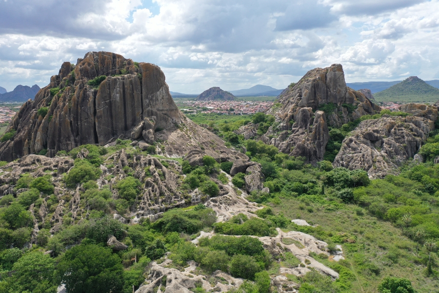 Fotografia aérea destaca duas imensas formações de rocha granítica escura e acinzentada que emergem verticalmente da paisagem. As rochas possuem texturas rugosas, com fendas profundas e marcas de erosão milenar, sendo parcialmente cobertas por uma vegetação rasteira e arbustos verdes que crescem em suas encostas e na base. Entre os dois grandes blocos de pedra, estende-se um vale de mata verde densa, e ao fundo, sob um céu claro com nuvens brancas e esparsas, avista-se o contorno de uma cidade pequena e outras silhuetas de montanhas distantes