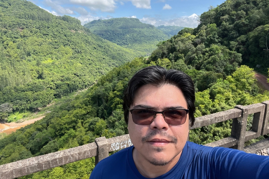 Foto de um homem em primeiro plano, com cabelos escuros e lisos, usando óculos escuros e camiseta azul, fotografando a si mesmo diante de uma paisagem natural exuberante. Ele está apoiado em um parapeito de concreto, possivelmente de uma ponte ou mirante. Ao fundo, estende-se um vale coberto por densa vegetação verde, com encostas de morros e montanhas sucessivas recobertas por floresta. Um curso d’água sinuoso aparece ao fundo, parcialmente visível entre as árvores, e o céu claro, com poucas nuvens