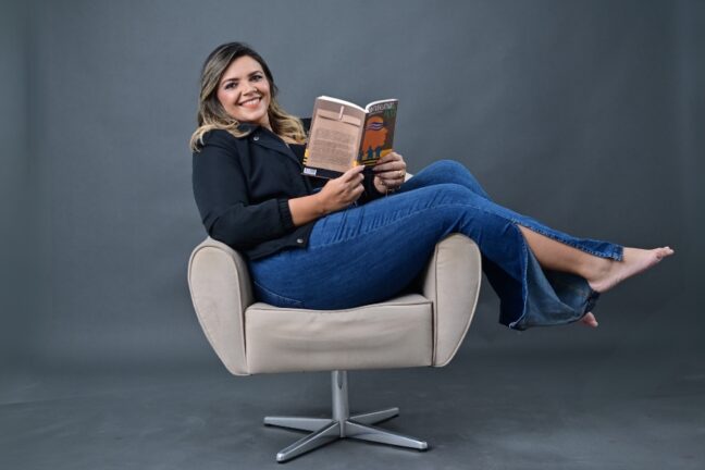 Uma mulher branca de pele clara e cabelos loiros ondulados sorri para a câmera enquanto posa sentada de forma descontraída em uma poltrona bege de base giratória metálica. Ela veste um blazer preto e calças jeans de corte "flare" com aberturas laterais nas barras, estando com os pés descalços apoiados no braço direito da poltrona. Com as mãos, ela segura um livro aberto, intitulado "Sustentabilidade 4.0", como se estivesse lendo no momento da foto. O fundo é de um cinza escuro uniforme e neutro