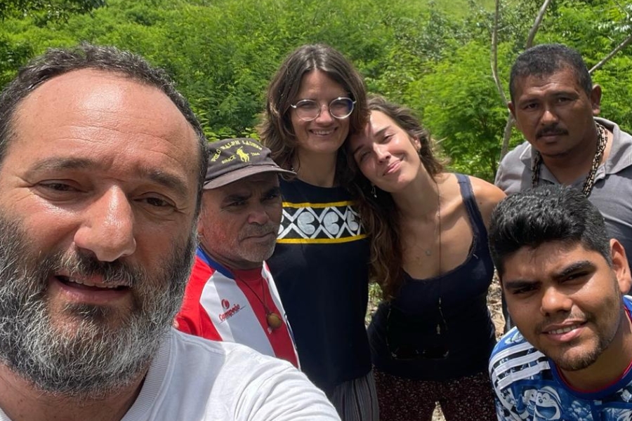 Selfie coletiva em meio à vegetação verde, mostrando homem em primeiro plano à esquerda, com barba grisalha e sorriso leve, acompanhado por outras pessoas, incluindo jovens e lideranças indígenas, todos sorridentes