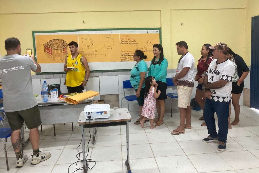 Em uma sala de aula, um rapaz pardo faz uma apresentação com desenhos técnicos e esquemas de arquitetura em um grande papel pardo fixado no quadro negro, enquanto um homem fotografa a cena e membros da sua equipe, incluindo adultos e uma criança, acompanham a explicação atentamente