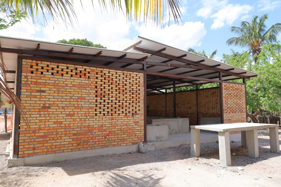 Uma estrutura moderna e funcional feita de tijolos aparentes com tramas vazadas para ventilação (cobogós), telhado metálico e bancadas de concreto, cercada por árvores e sob um céu aberto