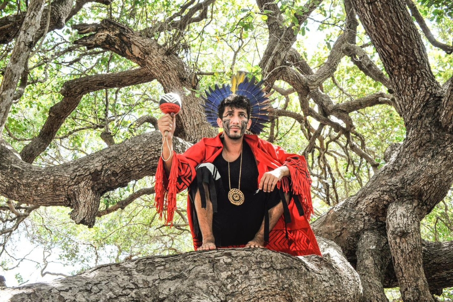 Jovem pardo agachado entre os troncos de uma árvore frondosa. Ele usa pinturas faciais étnicas, um cocar de penas azuis e amarelas, uma capa vermelha com franjas e segura um maracá