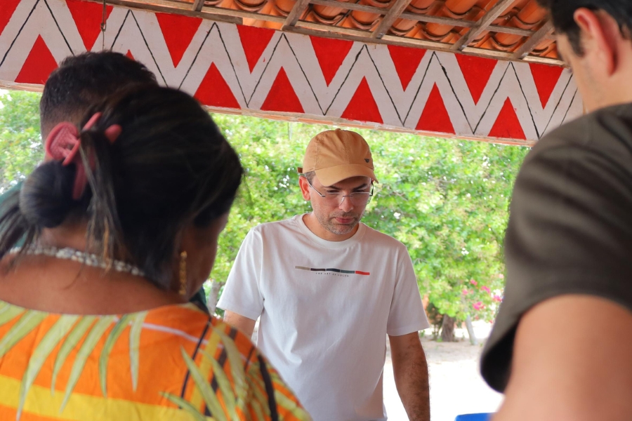 Homem branco de óculos e boné bege, vestindo uma camiseta branca, em um momento de interação com pessoas à sua frente (de costas na imagem) sob um teto decorado com padrões geométricos indígenas em vermelho e branco