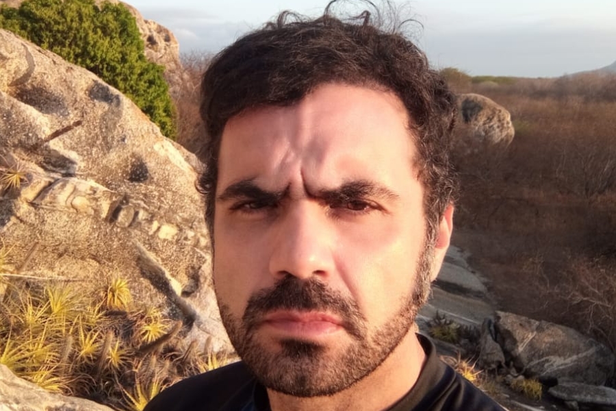 A foto mostra um homem de pele clara, cabelos escuros e levemente ondulados, barba e bigode bem aparados, enquadrado em primeiro plano, olhando diretamente para a câmera com expressão séria e concentrada. Ele veste uma camiseta escura. Ao fundo, há uma paisagem natural de aparência semiárida, com grandes formações rochosas, vegetação baixa e seca, além de algumas plantas espinhosas presas às rochas. A luz do sol, aparentemente no fim da tarde, ilumina o rosto e as pedras, criando tons quentes e sombras suaves, enquanto o horizonte revela um terreno irregular e árido, típico de áreas do interior nordestino