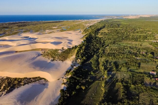 Do lado esquerdo, extensas dunas de areia branca e ondulada se estendem em direção ao oceano azul profundo no horizonte, com pequenas lagoas de água doce formadas entre os vales de areia. Do lado direito, uma vegetação densa e muito verde cria uma linha divisória nítida com as dunas, revelando áreas de mata preservada e pequenas propriedades rurais com plantações organizadas. Ao fundo, no canto superior direito, é possível ver uma fileira de aerogeradores brancos (turbinas eólicas) e o ponto onde um rio encontra o mar