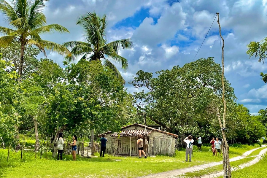 Uma fotografia ao ar livre, sob um céu azul com nuvens brancas, mostra um grupo de pessoas visitando uma casa tradicional de taipa em uma área rural e arborizada. A casa é pequena e rústica, com paredes de barro reforçadas por estacas de madeira e um telhado de telhas cerâmicas envelhecidas. O grupo, composto por cerca de oito pessoas em trajes casuais, está espalhado pelo terreno gramado: algumas observam a construção de perto, enquanto outras conversam ou fotografam a paisagem. Ao redor da casa, há uma vegetação exuberante com árvores frondosas e coqueiros altos, e à direita, uma estrada de terra branca com marcas de pneus desaparece na curva