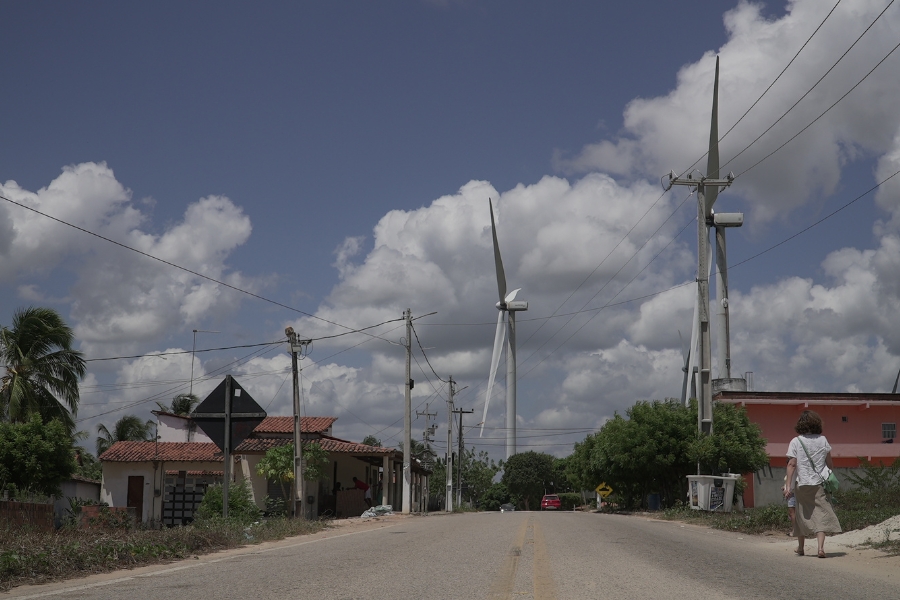 Fotografia de uma rua de asfalto onde a vida cotidiana se mistura com a infraestrutura de energia renovável. Em primeiro plano, uma estrada central com faixas amarelas duplas guia o olhar em direção ao horizonte, com uma mulher de costas caminhando pelo lado direito da via. O cenário é composto por casas simples de telhado de barro, postes de energia com fiação exposta e vegetação verde. O elemento mais marcante são as gigantescas torres de energia eólica brancas que se erguem logo atrás das moradias, com suas grandes pás dominando a paisagem sob um céu azul intenso e repleto de nuvens brancas volumosas