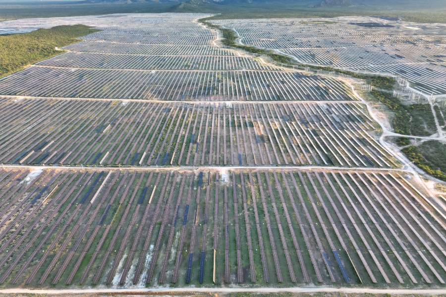 Esta fotografia captura uma rua de asfalto em Jurema, Amontada, no Ceará, onde a vida cotidiana se mistura com a infraestrutura de energia renovável. Em primeiro plano, uma estrada central com faixas amarelas duplas guia o olhar em direção ao horizonte, com uma mulher de costas caminhando pelo lado direito da via. O cenário é composto por casas simples de telhado de barro, postes de energia com fiação exposta e vegetação verde típica da região. O elemento mais marcante são as gigantescas torres de energia eólica brancas que se erguem logo atrás das moradias, com suas grandes pás dominando a paisagem sob um céu azul intenso e repleto de nuvens brancas volumosas.