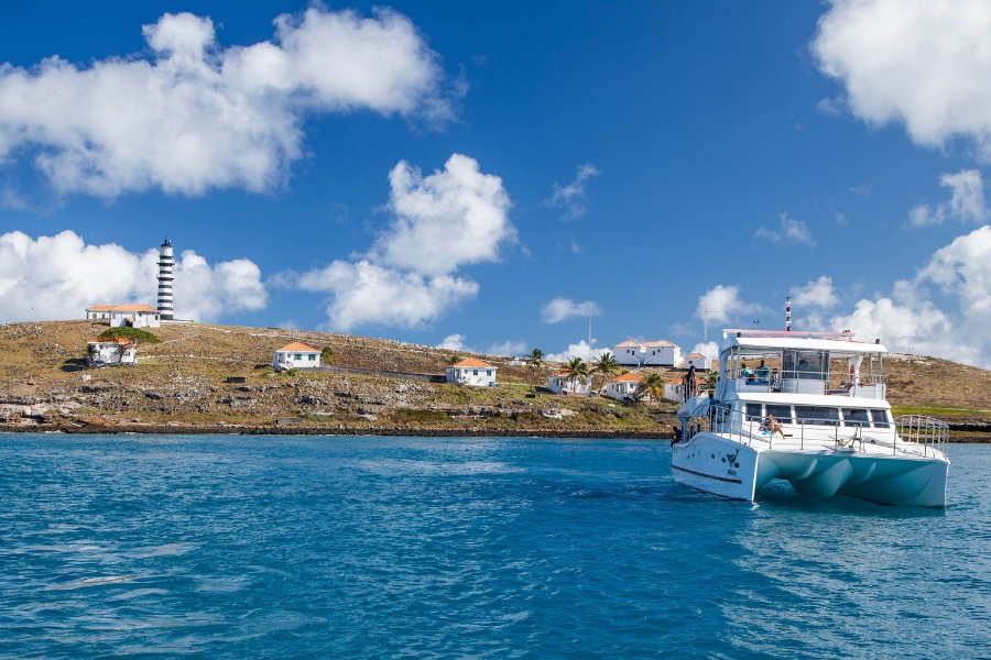 Um catamarã branco navega em águas azuis cristalinas sob céu ensolarado. Ao fundo, a Ilha de Santa Bárbara exibe o farol listrado e diversas casas brancas espalhadas pelo relevo árido.