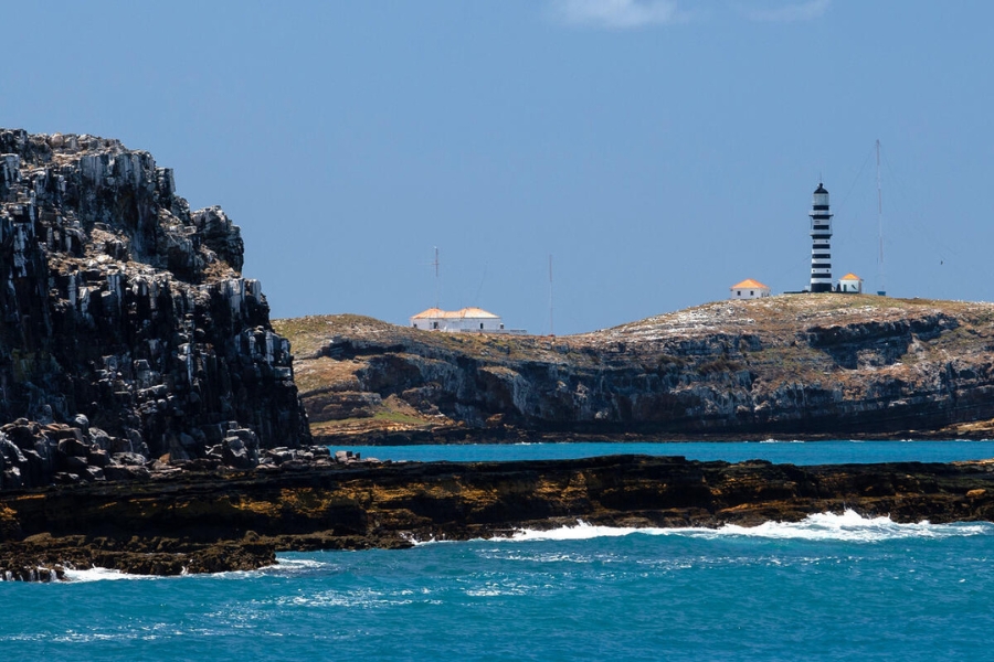 Vista ao nível do mar de encostas rochosas e escuras. No topo de uma colina ao fundo, avistam-se o farol preto e branco e pequenas construções sob um céu azul limpo.