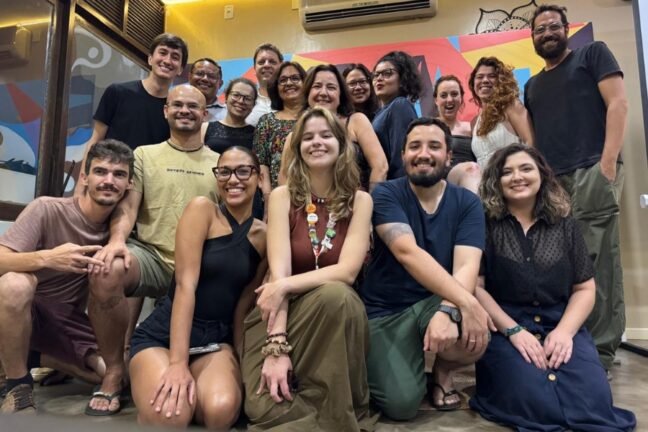 Uma fotografia em plano médio captura um grupo diverso de aproximadamente 15 pessoas reunidas em um ambiente interno iluminado, posando sorridentes para a câmera. O grupo está organizado em três níveis: alguns estão agachados na frente, outros sentados ao centro e o restante de pé ao fundo. No centro da imagem, em destaque, uma mulher jovem de cabelos loiros ondulados veste uma regata marrom e uma saia longa verde-oliva. Os integrantes vestem roupas casuais e variadas, incluindo camisetas, blusas de alças e vestidos em tons de preto, branco, azul e cores terrosas. O fundo é composto por uma parede amarela clara com um painel artístico colorido ao centro e uma porta de madeira com vidro à esquerda