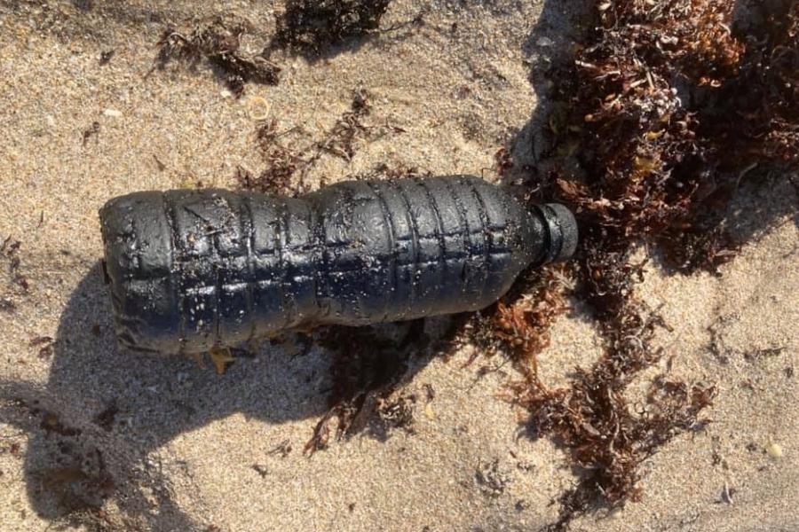 Foto em close mostra uma garrafa plástica descartada sobre a areia de uma praia, completamente revestida por uma camada espessa e fosca de óleo negro ou piche. A garrafa, que possui um formato cilíndrico com ondulações horizontais, está deitada horizontalmente e ocupa o centro da composição. Ao seu redor, a areia clara está misturada a pedaços de algas marinhas secas de cor castanho-escuro. Uma sombra nítida é projetada à esquerda da garrafa, indicando a incidência de luz solar lateral, o que ressalta a textura irregular e pegajosa do resíduo poluente que a envolve
