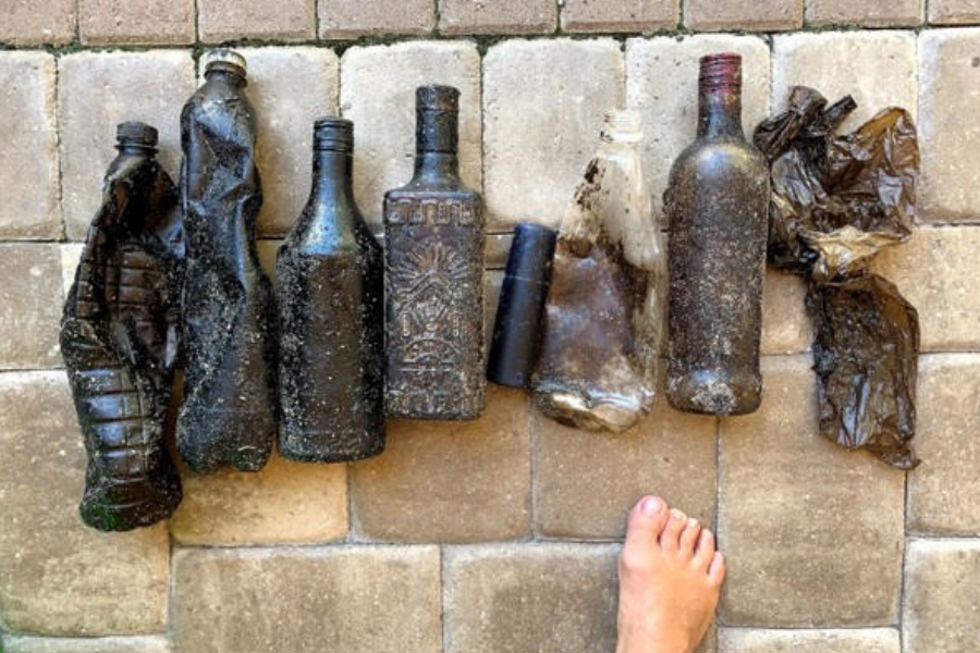 Foto apresenta uma fileira de seis garrafas e dois fragmentos de plástico dispostos verticalmente sobre um chão de pedras retangulares claras, evidenciando o impacto da poluição marinha. Da esquerda para a direita, as garrafas variam em material e estado: as quatro primeiras estão quase totalmente cobertas por uma substância preta e viscosa, como óleo ou piche, com algumas incrustações de areia; as outras duas são de plástico transparente, porém amassadas e sujas de resíduos escuros. À direita, há pedaços de plástico fino e enrugado, também manchados. Na parte inferior da foto, um pé humano descalço aparece parcialmente, servindo como escala para o tamanho dos objetos