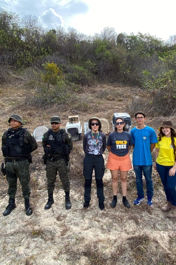 A imagem registra um grupo de seis pessoas posicionado lado a lado em um terreno arenoso e com vegetação baixa, tendo ao fundo uma colina repleta de arbustos sob um céu nublado. Da esquerda para a direita, os dois primeiros homens vestem fardamento militar camuflado completo, com coletes táticos e chapéus de aba curta; ao centro, uma mulher usa calças pretas, camiseta cinza e chapéu de trilha, seguida por outra mulher de camiseta cinza com a frase "WILD and FREE" e bermuda coral. À direita, um jovem veste camiseta azul e calça jeans, e uma jovem encerra a fila com camiseta amarela e calça azul. Atrás do grupo, sobre o solo, veem-se diversas caixas de transporte de animais e gaiolas, sugerindo uma ação de soltura ou resgate de fauna silvestre. Todos olham para a câmera em uma postura organizada, indicando uma parceria entre forças de segurança e civis em uma área de preservação.