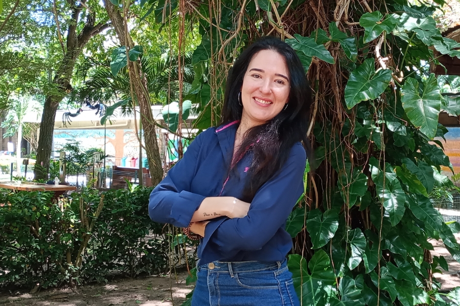 Uma mulher branca de longos cabelos pretos e sorridente posa no centro da imagem, com os braços cruzados sobre uma camisa social azul-marinho e calça jeans. Ela está em um ambiente externo arborizado, encostada em um tronco de árvore repleto de folhagens verdes e trepadeiras, com arbustos e luz solar filtrada ao fundo, transmitindo uma sensação de serenidade e simpatia