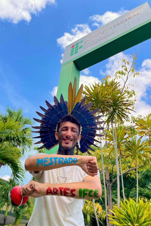 A imagem apresenta em ângulo baixo um homem jovem sorridente, de pele clara e barba curta, posando sob um totem verde do "INSTITUTO FEDERAL Ceará Campus Fortaleza". Ele veste um grande cocar de penas azuis e amarelas e tem o rosto pintado com grafismos indígenas pretos que se estendem das bochechas ao pescoço. Seus antebraços estão cruzados à frente do peito: no braço superior, lê-se a palavra "MESTRADO" escrita em azul; no inferior, "ARTES IFCE" em vermelho e verde. Com a mão direita, ele segura um maracá decorado em vermelho, preto e branco. Ele usa uma camiseta branca, colares de contas claras e o fundo é composto por palmeiras, folhagens tropicais e um céu azul com nuvens esparsas, evocando um momento de celebração e orgulho por uma conquista acadêmica.