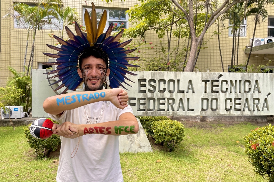 A imagem apresenta um homem jovem, de pele clara e barba curta, sorrindo de forma expressiva enquanto posa em frente a um letreiro de concreto onde se lê "ESCOLA TÉCNICA FEDERAL DO CEARÁ". Ele utiliza um imponente cocar indígena com penas em tons de azul vibrante, marrom e amarelo, além de pinturas gráficas pretas que descem de seus olhos pelas bochechas. O homem segura um maracá indígena decorado na mão direita e posiciona os antebraços cruzados à frente do corpo; neles, estão escritas as palavras "MESTRADO" em azul no braço superior e "ARTES IFCE" em vermelho e verde no braço inferior. Ele veste uma camiseta branca e colares de contas claras. O cenário é uma área externa gramada, com arbustos e árvores ao fundo, sob a luz do dia, transmitindo um sentimento de celebração e conquista acadêmica.