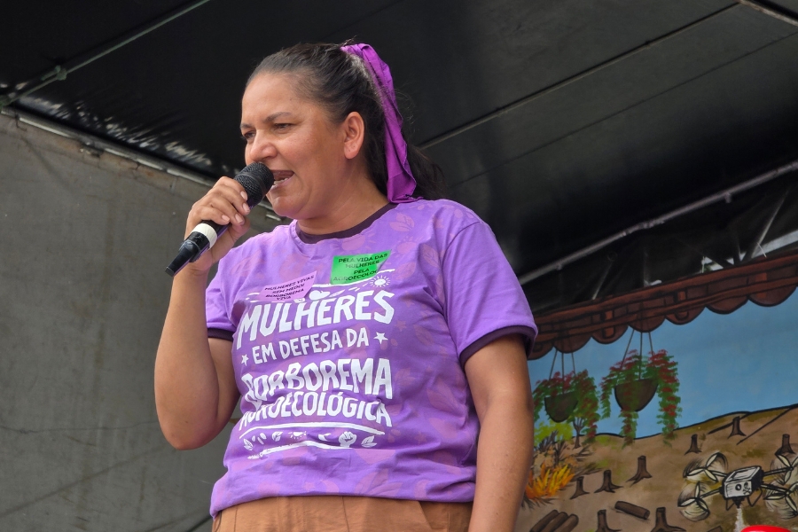Mulher retratada em plano médio, falando ao microfone com um semblante sério e decidido. Ela é parda, de cabelos pretos presos com um lenço roxo, e veste a camiseta lilás da marcha com os dizeres "Mulheres em defesa da Borborema Agroecológica". Está posicionada em um palco, sob um teto escuro. Ao fundo, à direita, observa-se parte de um painel ilustrado que retrata cenários de degradação ambiental, como queimadas e o uso de drones em áreas desmatadas