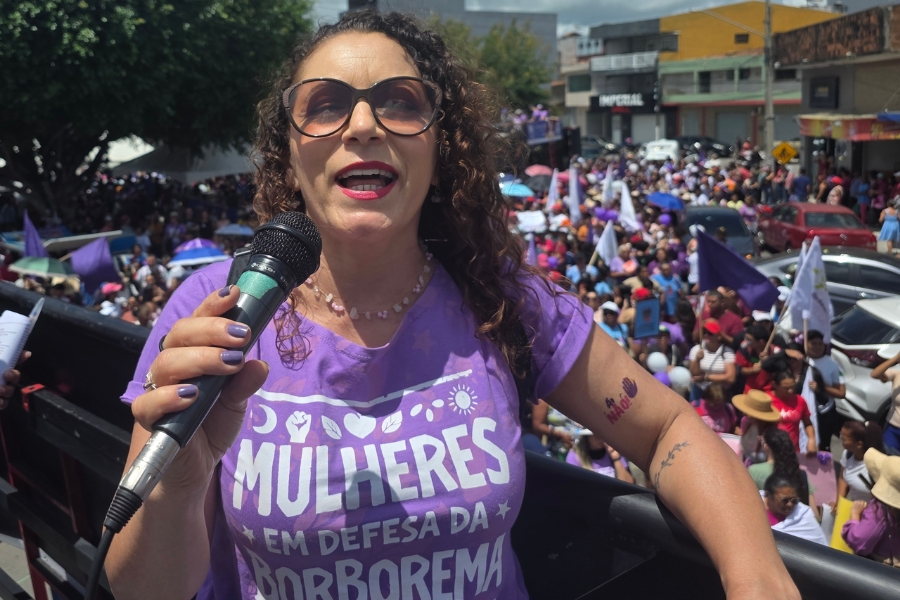 Em primeiro plano, uma mulher branca de cabelos castanhos cacheados e longos, usa óculos de sol grandes e redondos e segura um microfone preto, falando com expressão entusiasmada. Ela veste a camiseta lilás onde se lê "Mulheres em defesa da Borborema", e tem uma pequena tatuagem no braço esquerdo com o símbolo de uma mão e a palavra "Não". Ela está posicionada no alto de um carro de som, cujas barras pretas aparecem na parte inferior da imagem. Ao fundo, a rua está tomada por uma multidão de pessoas, bandeiras roxas e carros, sob a luz intensa do sol