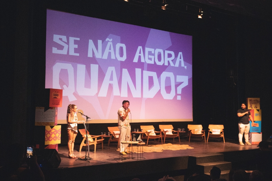 A imagem captura o palco de um auditório durante um evento institucional. Ao centro, um homem de camisa clara fala ao microfone, ladeado por uma mulher à esquerda, que consulta papéis em um pedestal, e um intérprete de Libras à direita. Ao fundo, uma grande tela exibe a frase em português "SE NÃO AGORA, QUANDO?" em letras garrafais brancas sobre um fundo em tons de roxo e lilás. O cenário é composto por cinco poltronas de madeira dispostas em semicírculo e totens coloridos com ilustrações e textos nas laterais do palco. A iluminação é focada nos participantes, contrastando com a penumbra da plateia, sugerindo um ambiente de debate ou conferência formal.