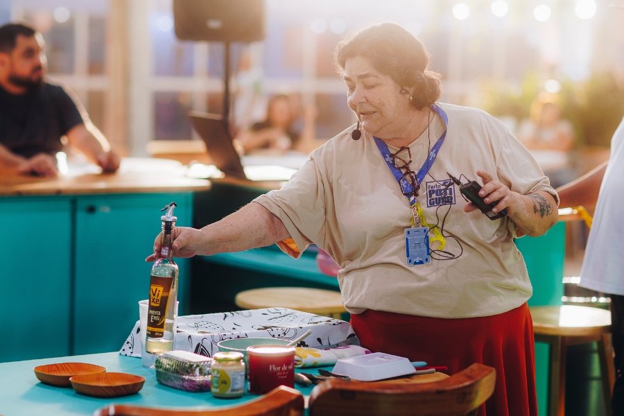 A imagem mostra uma mulher de pele clara e cabelos escuros presos, em um ambiente de oficina ou cozinha experimental. Ela veste uma camiseta bege com a logo "Feito no Potiguar", uma saia vermelha, usa óculos pendurados no decote e um microfone auricular, indicando que está realizando uma demonstração. Com a mão direita, ela alcança uma garrafa de vidro transparente sobre uma mesa azul, onde também estão organizados pequenos bowls de madeira, potes de condimentos e uma embalagem de "Flor de Sal". O fundo é amplo e iluminado com luzes quentes, apresentando um balcão azul e outras pessoas de forma desfocada, o que confere um aspecto profissional e dinâmico à cena.