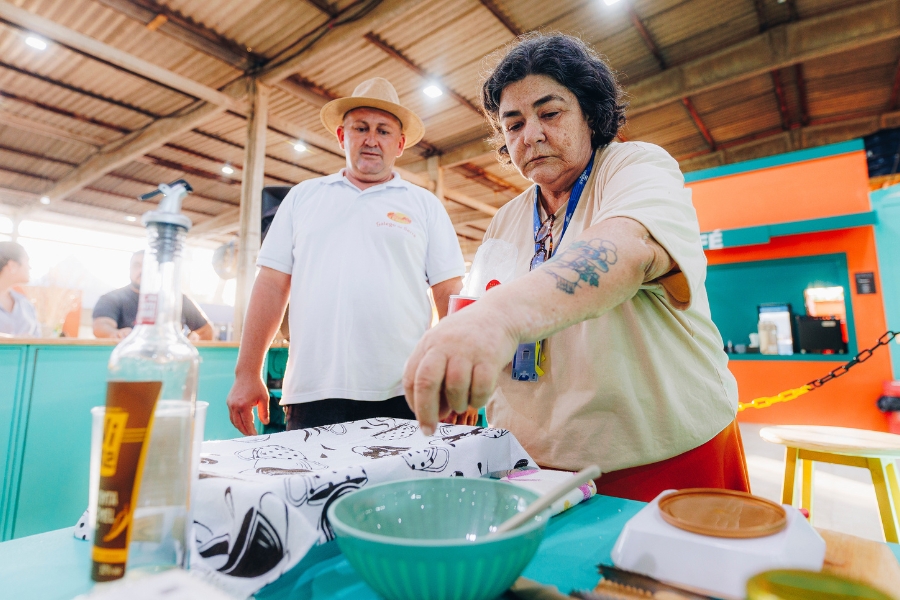 Nesta imagem, a mulher de pele clara e cabelos escuros, agora vista de um ângulo mais baixo e próximo, aparece concentrada enquanto tempera ou finaliza um preparo. Ela estende a mão direita sobre uma tigela de cerâmica verde-água que está em uma mesa azul, enquanto a mão esquerda, que possui uma tatuagem detalhada no antebraço, segura um recipiente. Ao seu lado, um homem de pele clara, vestindo uma camisa polo branca e chapéu de palha, observa a ação com atenção. O cenário é um amplo galpão com teto de telhas metálicas e iluminação industrial; ao fundo, destaca-se uma estrutura laranja vibrante e balcões em tons de azul, reforçando o ambiente de um evento gastronômico ou feira cultural.